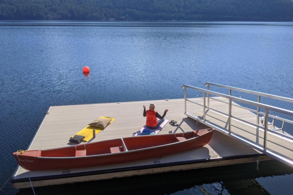 Home Woman practicing yoga and meditation on the private dock at Christina Lake House next to a red canoe.